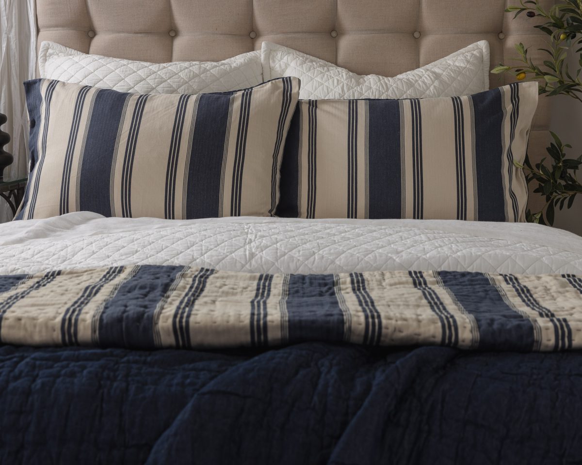Amity home camden quilt   blue and beige ticking stripe styled on a bed with a navy quilt and matching striped pillows
