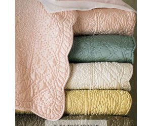 Amity home tudor quilt   pink with intricate quilted medallion pattern and scalloped edge draped over a stack of luxury quilts in various colors